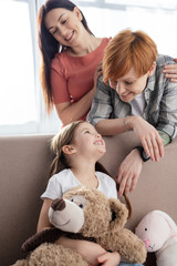 Selective focus of cheerful kid with soft toys on couch looking at same sex parents in living room
