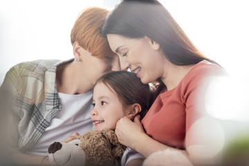Selective focus of happy same sex parents kissing and hugging smiling daughter with teddy bear isolated on white