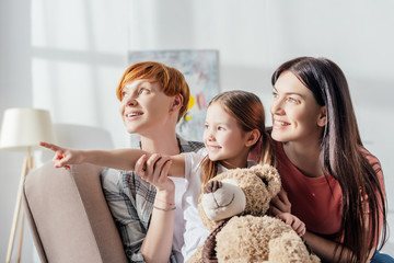Smiling kid with teddy bear pointing with finger near mothers while sitting on couch in living room