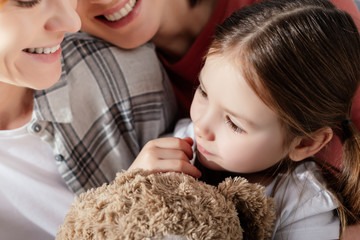 Child with teddy bear looking away near smiling same sex parents