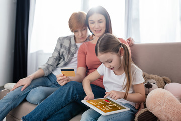 Child using digital tablet with e-health app near smiling same sex parents with credit card on couch