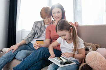 Kid using digital tablet with tickets app on screen near same sex parents with credit card at home