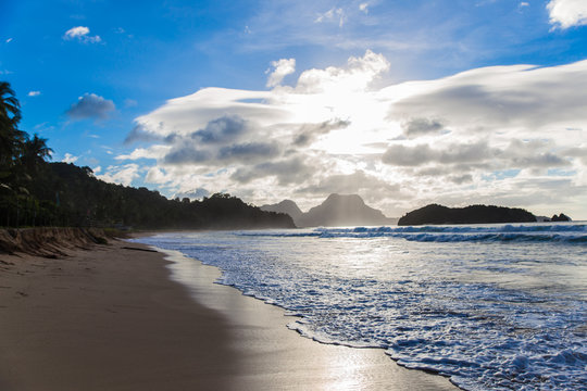 empty sand beach with waves and sun covered with cloudy sky and tropical trees on background in El Nido, Palawan province, Philippines