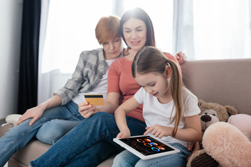 Same sex parents with credit card sitting near daughter using digital tablet with trading rate app