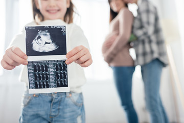 Cropped view of smiling child showing ultrasound scan of baby while woman embracing pregnant...