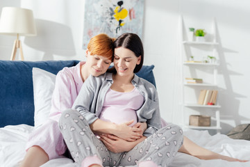 Smiling woman embracing pregnant girlfriend on bed