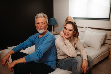 Obraz premium Portrait of elderly man and his brunette daughter sitting on bed and looking at camera. Blonde woman is talking over the phone while lying in bed behind them.