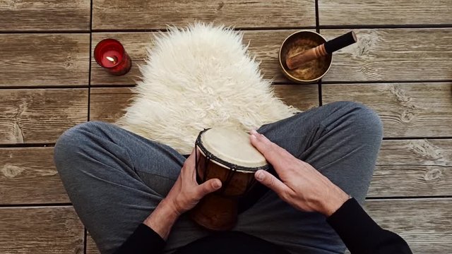 Uomo che medita suonando un piccolo bongo seduto a gambe incrociate su pavimento in legno