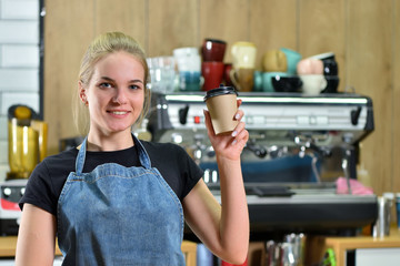 A young bartender girl in a jeans apron on a background of a coffee machine offers coffee to go. Concept of sale and work in a coffee shop in a small business.