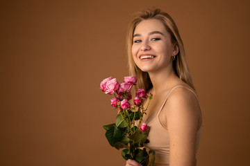 Cute young blonde haired girl in beige top, holding a bouquet of pink flowers, enjoying the smell, broadly smiling and looking at the camera, standing over dark orange background