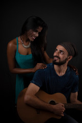 A dark skinned Indian/African girl and a Kashmiri/European/Arabian man in casual wear with guitar in front of a black copy space studio background. Indian lifestyle and fashion photography.