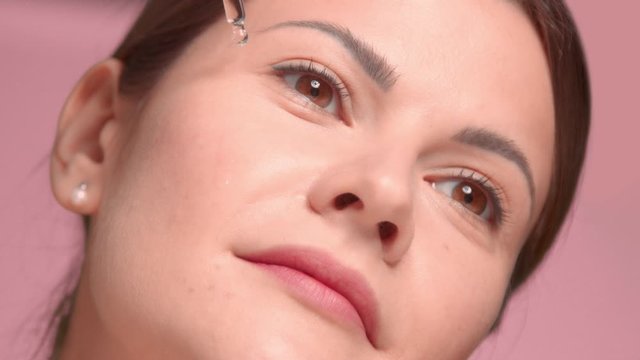 30s brunette woman closeup with an oil dropper put some oil on her cheek on pink background