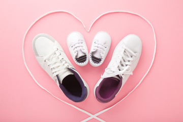Father, mother and little kid shoes. Heart created from white shoelaces. Light pink floor background. Pastel color. New family. Love sport together concept. Closeup. Top down view.