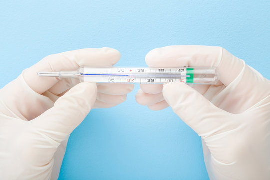 Doctor hands in white rubber protective gloves holding mercury thermometer on light blue table background. Pastel color. Closeup. Point of view shot. Top down view.