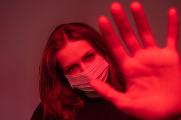 Portrait of infected young woman in face mask making stop gesture to camera, red light effect