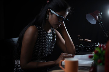 Young brunette Indian/African dark skinned lady in western dress and spectacles studying in front of a table lamp in black copy space studio background. Lifestyle and fashion.