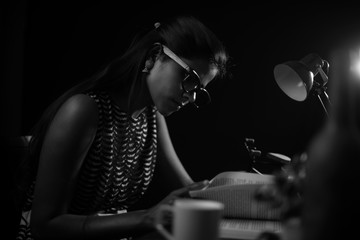 Young brunette Indian/African dark skinned lady in western dress and spectacles studying in front of a table lamp in black copy space studio background. Lifestyle and fashion.