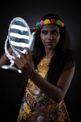 Fashion portrait of young dark skinned Indian Bengali brunette girl in floral dress and head band holding  mirror in front of black copy space studio background. Indian fashion portrait and lifestyle.