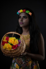 Fashion portrait of young dark skinned Indian Bengali brunette girl in floral dress with floral head band taking flowers in front of black copy space studio background. Indian fashion photography.