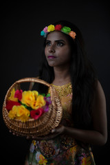 Fashion portrait of young dark skinned Indian Bengali brunette girl in floral dress with floral head band taking flowers in front of black copy space studio background. Indian fashion photography.