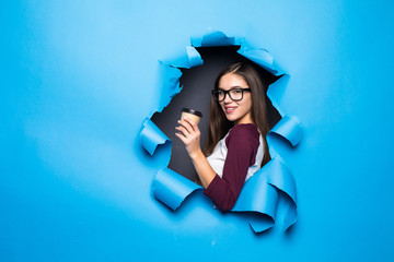 Young pretty woman holding coffee while looking through blue hole in paper wall.