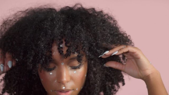 Mixed race black woman with curly hair covered by crystal makeup on pink background in studio lift up her hair from face