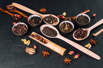 Assorted dried tea leaves in bowls and wooden spoons on a concrete black background. Dark moody. Set of different herbal and fruit tea