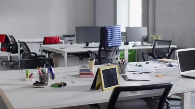 Tracking shot of empty open space office with modern and comfortable workplaces. There are desks with laptop, tablet, computers and stationery on them