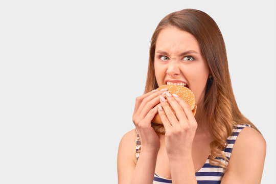Cute girl quickly eats Burger, wrinkling nose, getting angry, Baring teeth. Wears striped t-shirt, isolated on white background in Studio with space for advertising