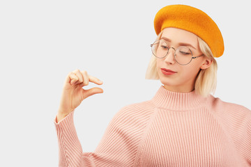 Smiling girl shows little amount of something, gestures with hand, being in high spirit. dressed in yellow beret and pink sweater, round big glasses, red lipstick. Isolated on white wall in Studio.