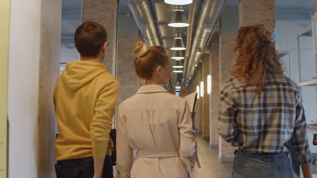 Rear view following shot of young multiethnic coworkers in casual outfits discussing something while walking through hallway of modern office building; their male colleague riding scooter