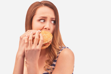 Cute girl quickly eats Burger, is afraid of being caught, hides from prying eyes. Wears striped t-shirt, isolated on white background in Studio with space for advertising