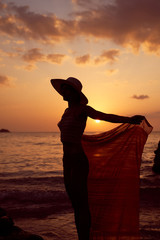 Fototapeta premium Contrast silhouette young slender woman against background of Sunny sunset, sea sandy beach. Warm evening tones. Lady walking on beach in sunset, vacation city Patong Phuket country Thailand, vertical