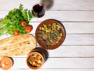 flat lay on plate and clay pot with georgian food chanakhi with beef and vegetables, bread lavash, parsley, red wine, tomato on white wooden background