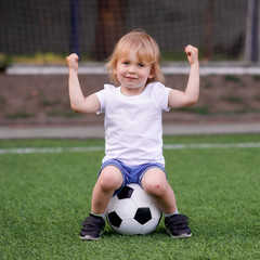 Little goalkeeper: blonde toddler kid sitting on soccer ball at green football field with arms raised, jokingly showing muscles and power. Success, achievement and gym class at kindergarten concept