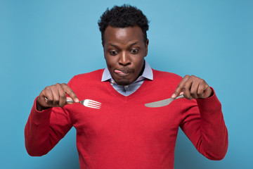 Young hungry crazy african american man in red sweater holding a fork and a knife. Isolated on blue...