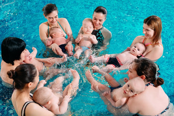 A group of mothers with their young children in a children's swimming class with a coach.