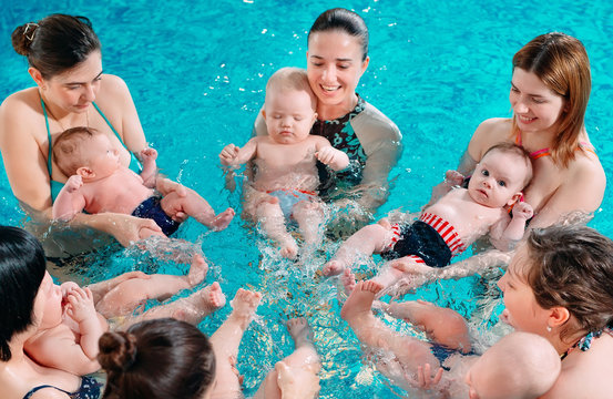 A group of mothers with their young children in a children's swimming class with a coach.