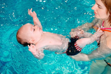 A group of mothers with their young children in a children's swimming class with a coach.