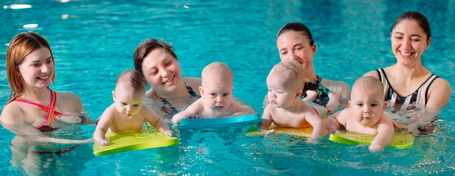 A group of mothers with their young children in a children's swimming class with a coach.