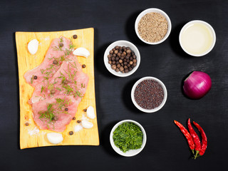 Sliced pieces of meat turkey game filet on a wooden cutting board, garlic, cumin (jeera), salt, fennel, allspice, mustard seeds, oil, red onion, red chili pepper on black wooden background. 