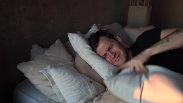 Young man sleeping on pillows in bed in early morning. Angry male covering his head with pillows because of noise that interferes with sleep.