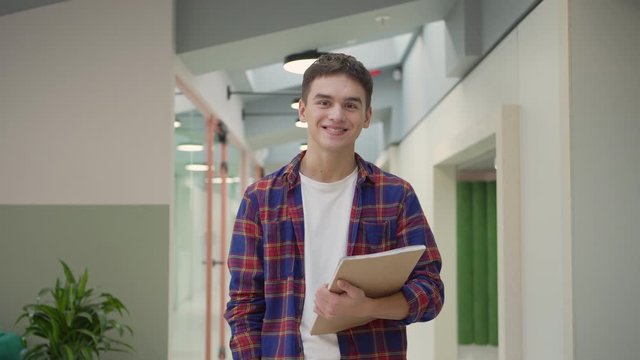 Tracking waist up portrait of smiling young man in checkered shirt looking at camera, smiling and nodding friendly while posing with notepad in college or office corridor