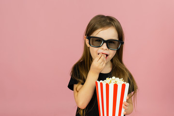 Fototapeta premium Cute little girl eats popcorn from striped packaging and watches a movie with 3D glasses. Cinema advertising concept, watching online movie