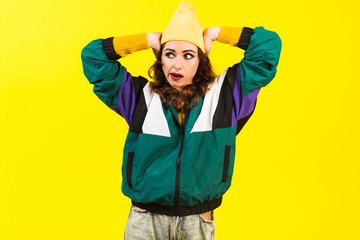 Modern beautiful girl in oversized sports jacket, hat and jeans, Billie Eilish style, back in the 90s, 2000s. Posing in the studio on a yellow background, strong funny emotions
