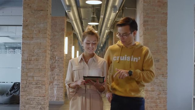 Young Caucasian Woman And Asian Man In Casual Clothes Smiling And Discussing Project On Digital Tablet While Walking Together Towards The Camera Through Loft Hallway