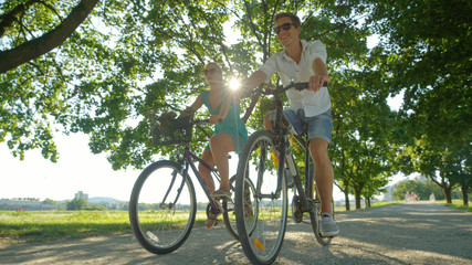 LOW ANGLE: Sun rays shine on happy tourist couple riding bicycles around a park