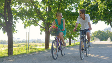 Young Caucasian couple smiles while riding bicycles down a scenic green avenue.