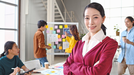 Portrait of beautiful Asian woman smiling, arms crossed in modern office, with young creative team work together. Female leader, company CEO manager, corporate organization, or business owner concept