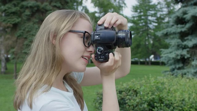 Attractive blonge young female-photographer in glasses takes picture in park and carefully evaluates the result before making next photo. Side view.